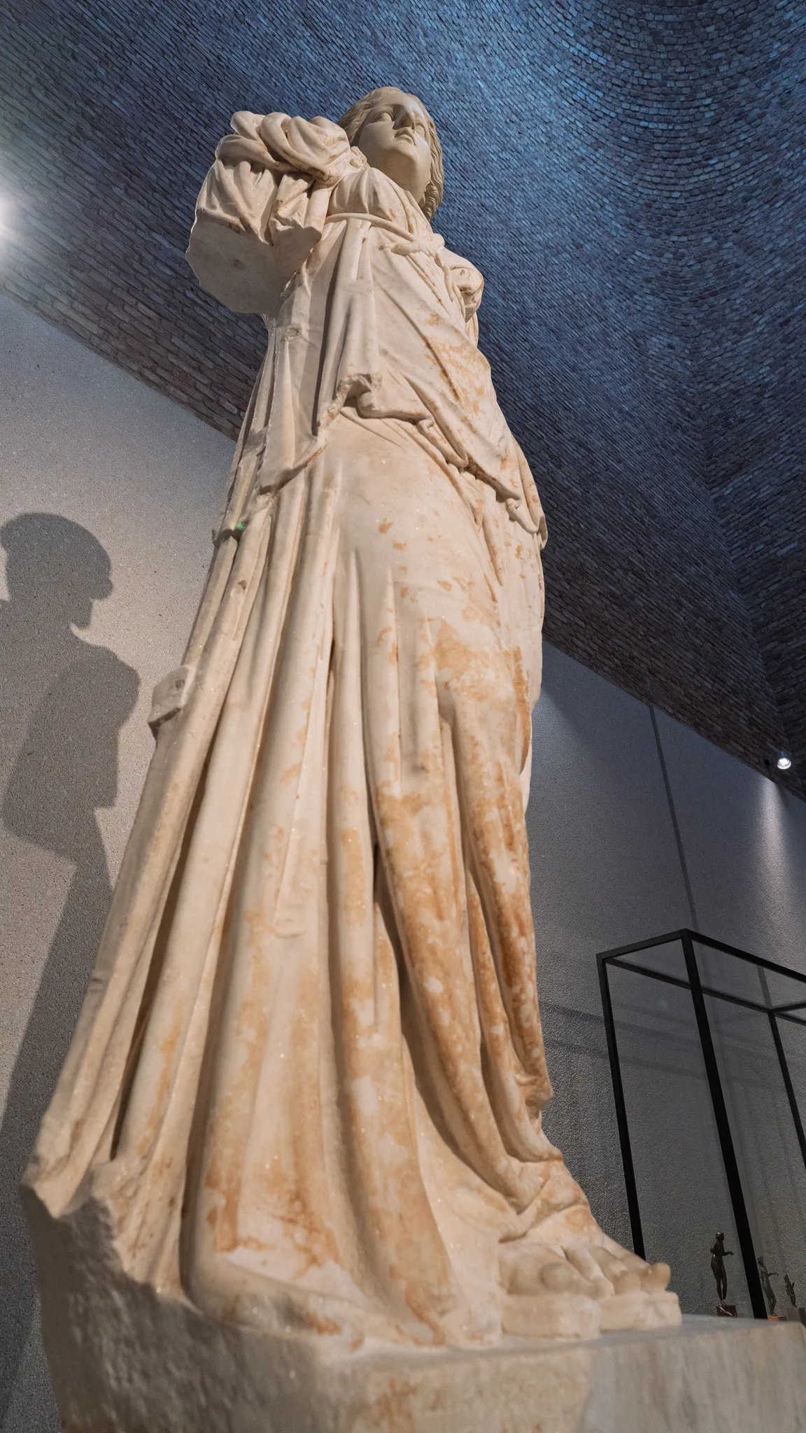 A weathered marble statue of a draped figure in a museum gallery