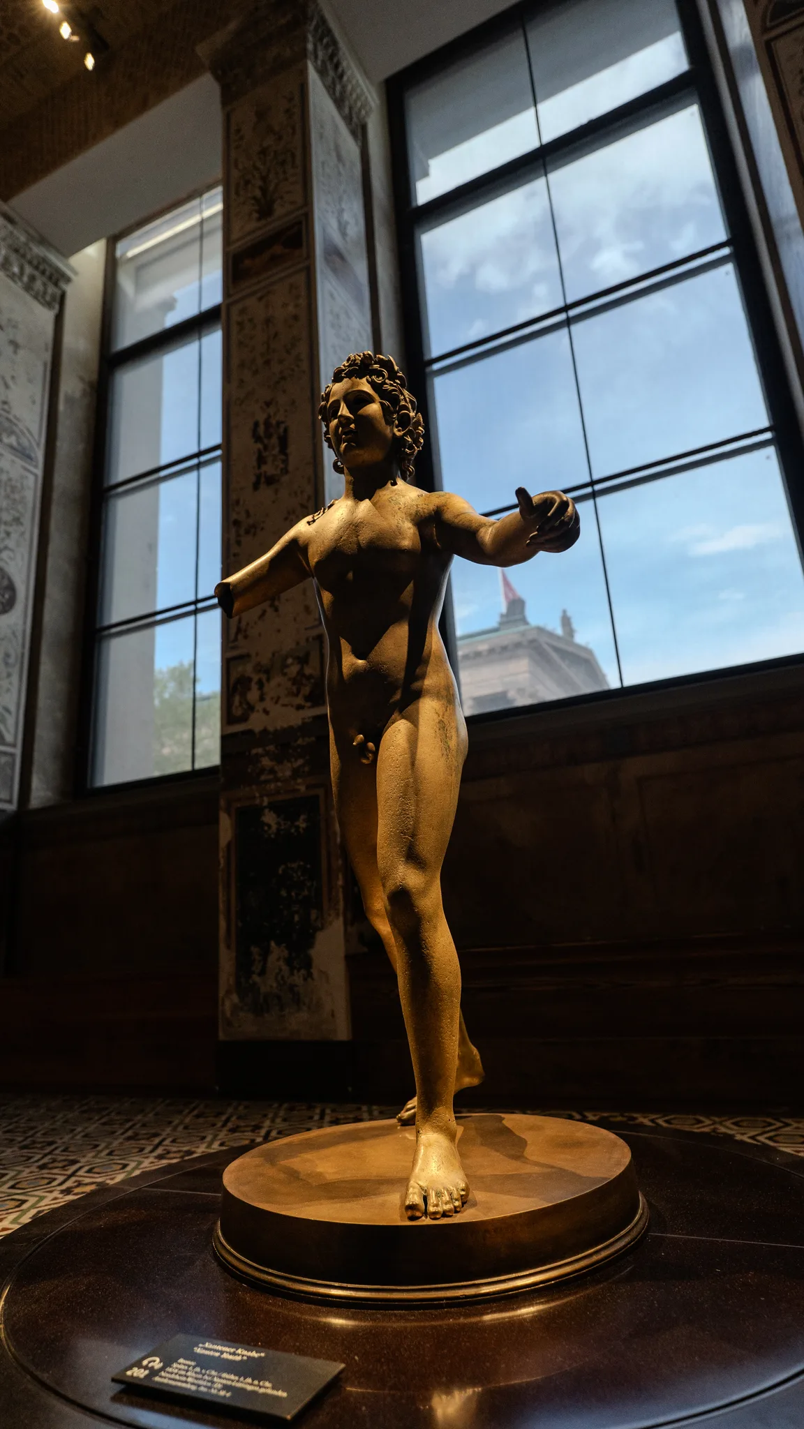 A small bronze statue of a young man with outstretched arms in a museum room with tall windows