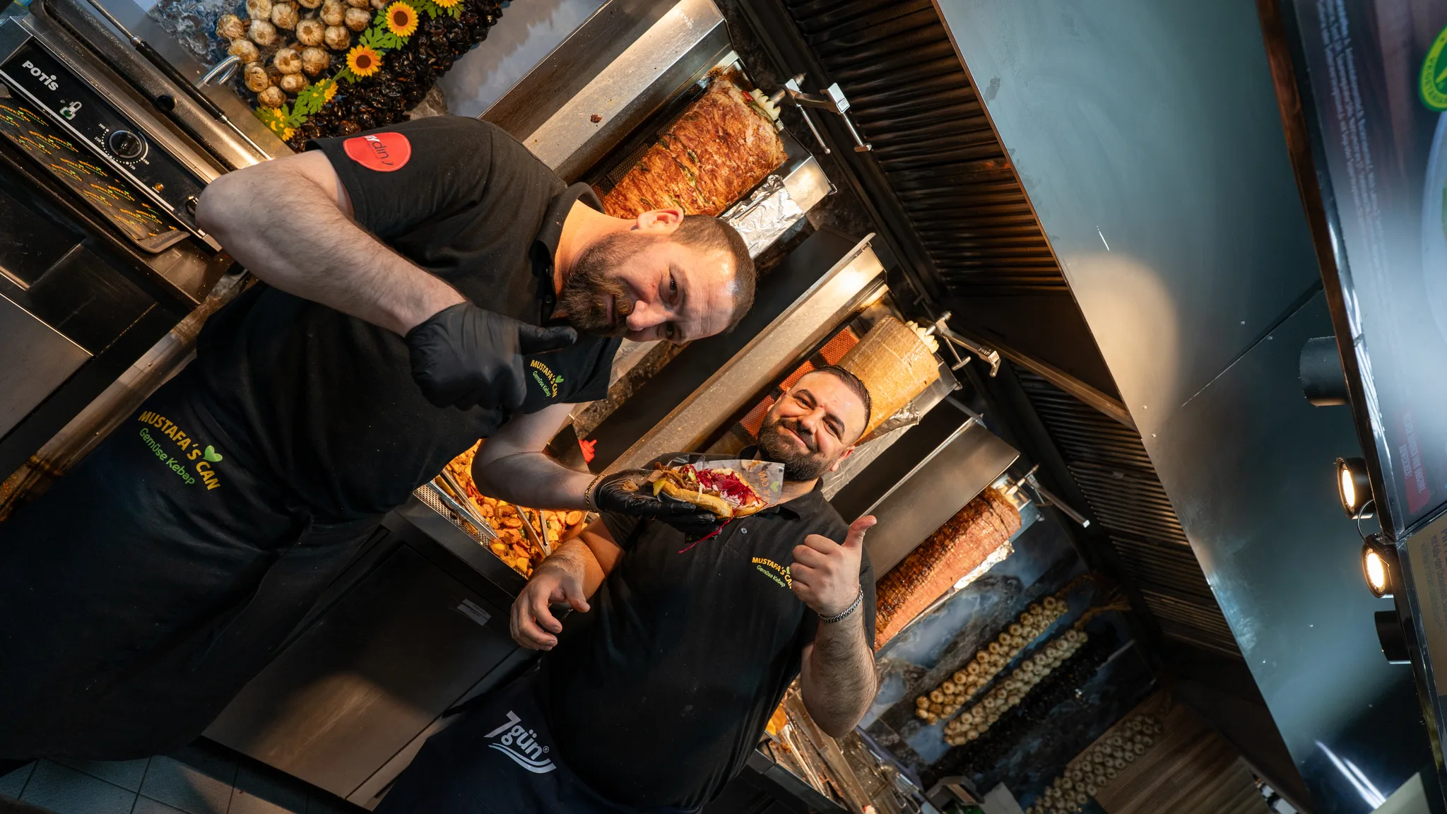 Two cooks at a busy kebab stall smiling at the camera, one holding up a kebab