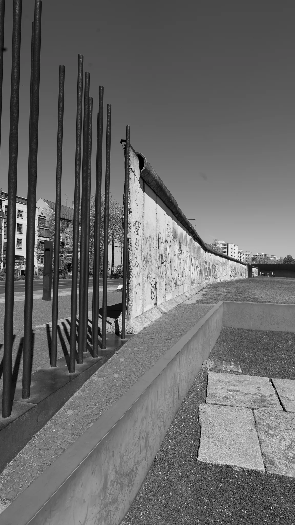 Black-and-white photo of a surviving section of the Berlin Wall beside a row of steel rods